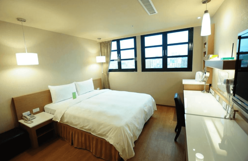 【高雄親子飯店】康橋商旅 高雄站前館的雙人房，採用米色牆面與木質家具設計，營造舒適氛圍。房內配有一張大床，白色床單整潔柔軟。窗邊設有書桌與工作區，提供充足照明，適合商務或旅遊住宿使用。
