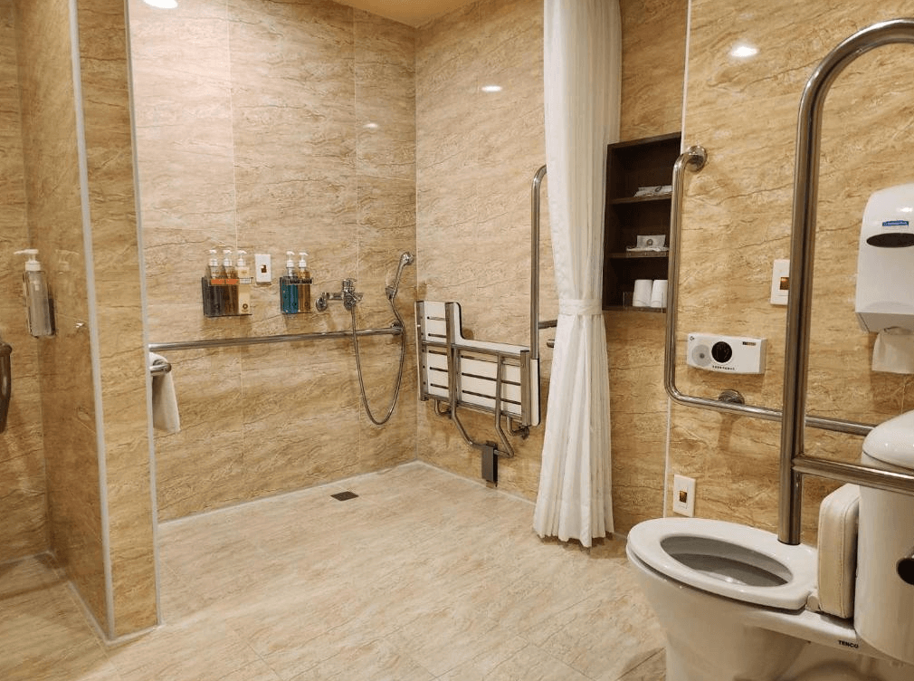 【高雄親子飯店】康橋商旅 高雄站前館的無障礙衛浴空間，採用淺色大理石瓷磚，展現乾淨明亮的環境。浴室內配有安全扶手、折疊椅與無障礙設施，方便行動不便的旅客使用。盥洗用品齊全，提供淋浴與座式馬桶，提升便利性。