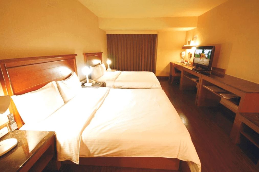 【員林住宿】康橋商旅-員林館的客房內部，設有兩張大床，木質床架搭配白色床單，整體風格溫暖且經典。床頭有檯燈，提供柔和照明，房內設有長書桌與壁掛電視，窗戶搭配深色窗簾，氛圍溫馨適合家庭或多人入住。