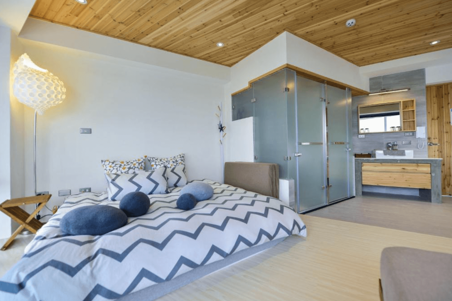 【台南安平住宿】思慕嶼 微光旅行的木質風格雙人房，天花板與浴室櫃體皆採原木色設計，搭配霧面玻璃隔間與簡約的灰白色調，床上擺放幾顆仿石頭造型抱枕，呈現自然舒適的住宿氛圍。