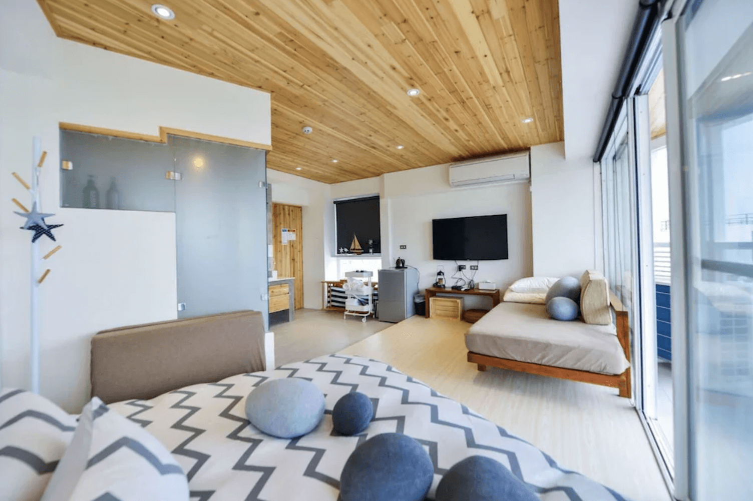 【台南安平住宿】思慕嶼 微光旅行採光極佳的開放式客房，木質天花搭配大面落地窗與玻璃拉門，室內設有沙發床、小冰箱與電視設備，整體設計溫暖明亮，適合親子或情侶住宿。