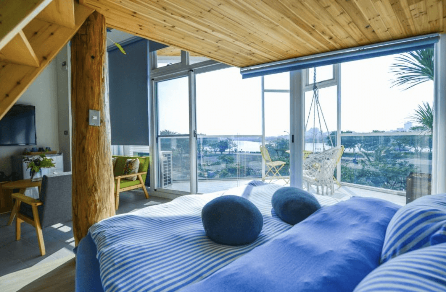 【台南安平住宿】思慕嶼 微光旅行的海景雙人房內部，大片落地窗可遠眺海灣風光，陽台設有吊椅與戶外座椅，床鋪以藍白條紋床包營造海洋風情，天花與樑柱為天然原木材質，充滿渡假氛圍。