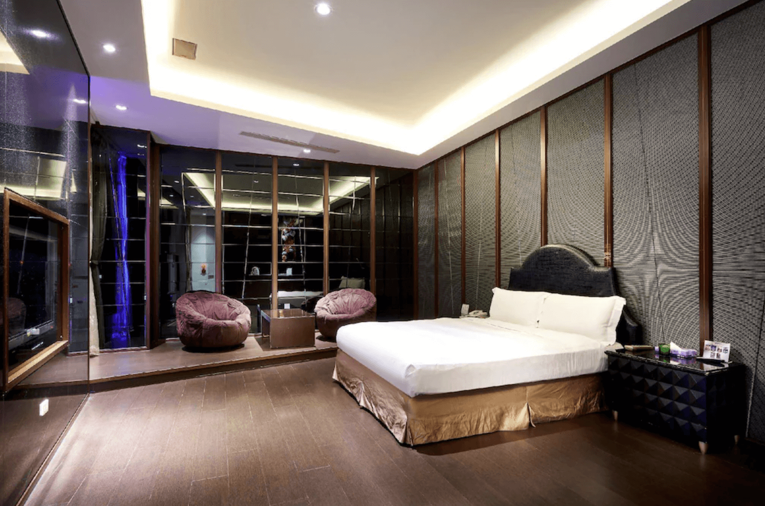 【小港住宿】打狗戀館的雙人房，房內設有一張大床與黑色絨面床頭板，牆面以深色線條紋理裝飾，搭配間接照明增添氛圍。床前有一小階平台區域，擺放兩張舒適的懶人椅與玻璃茶几，整體設計低調奢華，適合情侶入住。