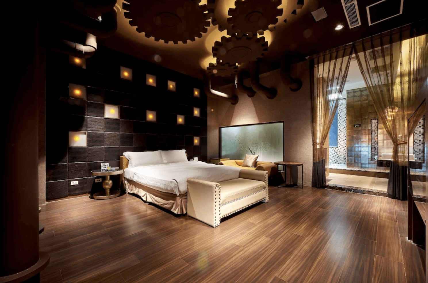 【小港住宿】打狗戀館以工業風結合蒸氣龐克風格設計的主題套房。天花板裝飾有大型齒輪造型，牆面為黑磚搭配暖黃燈光，營造出神秘浪漫氛圍。房內配有大床、沙發、工作桌與拉簾區隔的浴室空間，極具視覺張力與特色。