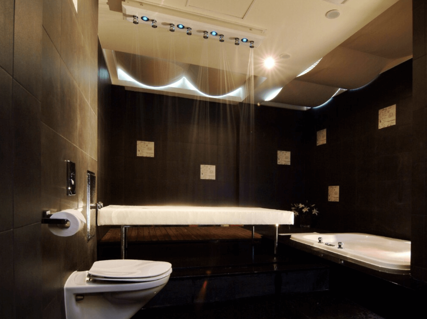 【小港住宿】打狗戀館內的專屬的浴室設施，整體以深色磁磚打造出沉穩氛圍。室內配有沖洗床、按摩浴缸與馬桶，並設有多組天花板噴頭裝置，可體驗SPA式淋浴。燈光設計柔和，適合放鬆與私人享受。