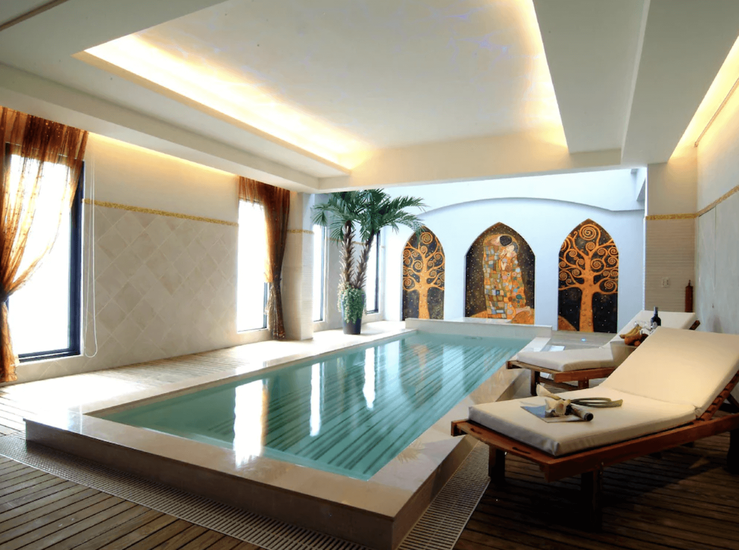 【小港住宿】打狗戀館的室內泳池空間，擁有獨立長型溫水池與舒適躺椅區，牆面繪有金色馬賽克風格壁畫，增添藝術氣息。自然光從大片窗戶灑落，搭配室內植栽與木質地板，營造出宛如度假村般的南洋風情。
