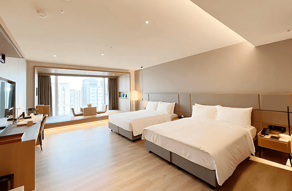【高雄親子飯店】承億酒店寬敞明亮的雙床房，設計風格簡約自然，以溫暖木質調與淺色系牆面為主。大片落地窗讓自然光灑入，窗邊設有和式座椅與小茶几，營造放鬆氛圍。房內配置書桌、電視與充足的收納空間，適合家庭或多人入住。