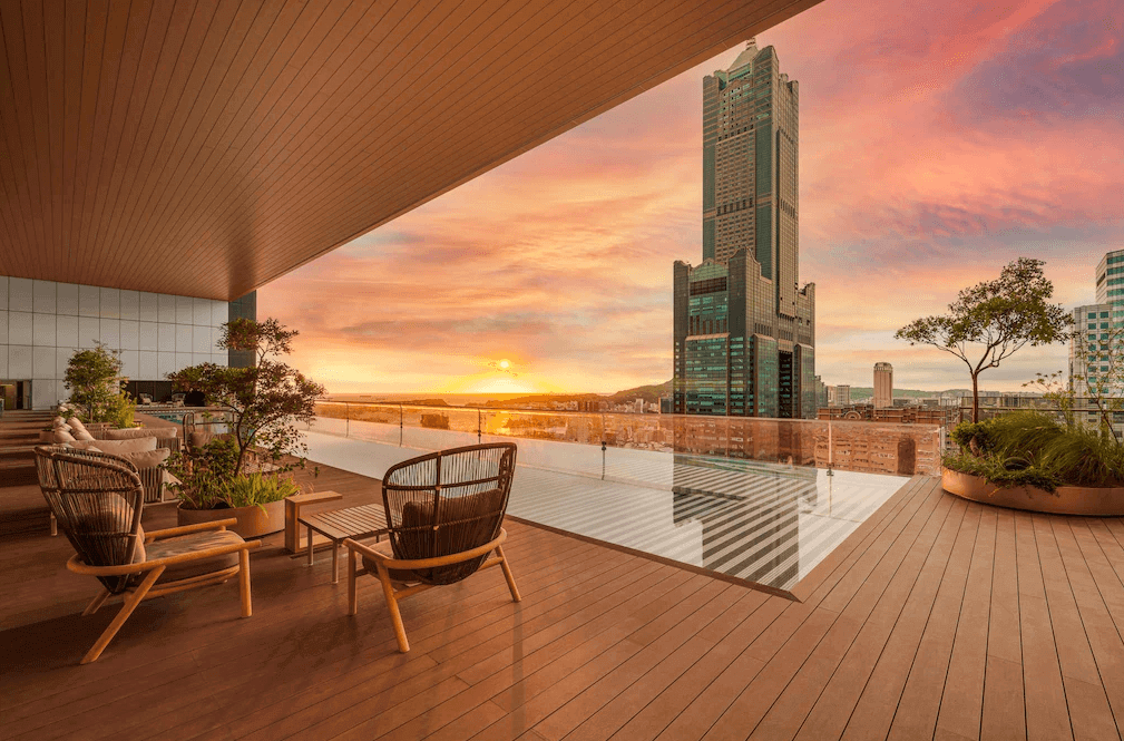 【高雄親子飯店】承億酒店的露天景觀陽台，設有舒適的藤編座椅與綠植點綴，提供休閒放鬆的空間。夕陽西下時，天空呈現夢幻般的粉橘色，遠處可見高雄著名的 85 大樓與城市天際線，景色壯麗迷人。