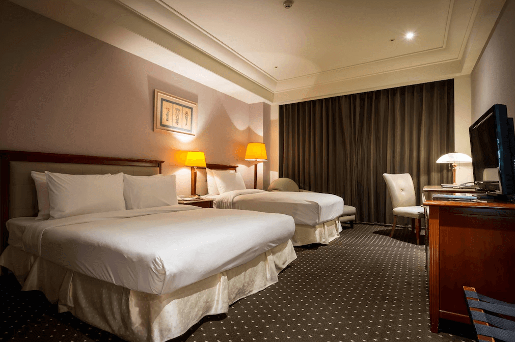【員林住宿】"彰化飯店"昇財麗禧酒店的高級客房，內有兩張大床，鋪設白色床單與典雅床裙，搭配柔和黃色燈光與木質家具，營造溫馨氛圍。房間設有厚重窗簾、書桌、電視與舒適座椅，適合家庭或商務旅客入住。