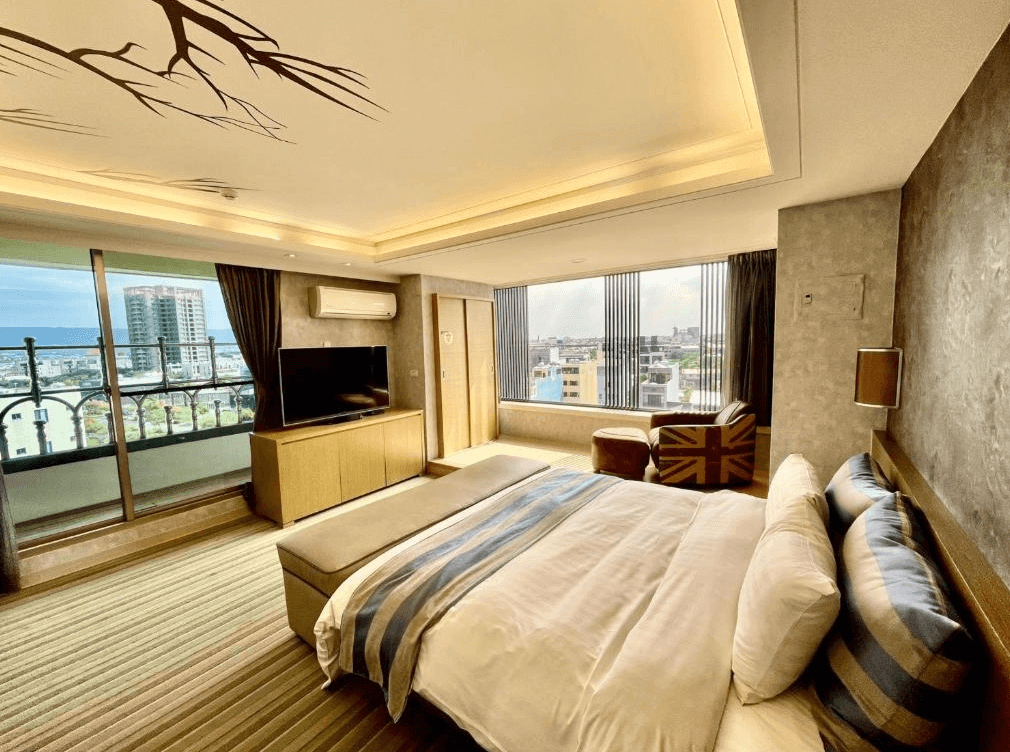 【員林住宿】晨陽文旅的高樓層套房，擁有大面落地窗與陽台，可遠眺城市與海景。房內設有一張大床，搭配藍白條紋床單，並擺放英國國旗圖案的單人椅，增添異國風情。天花板採用樹枝剪影裝飾，營造自然感。