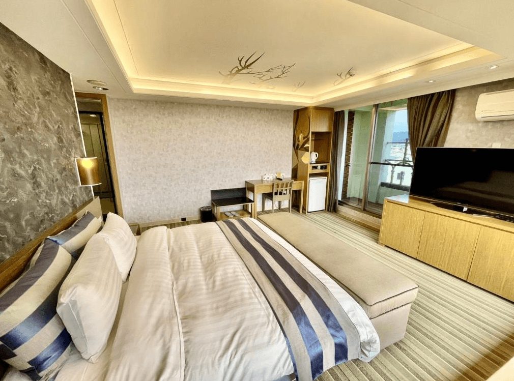 【員林住宿】晨陽文旅的飯店客房，設有一張大床與長型貴妃椅，床頭牆面為石紋質感設計。房內配有書桌、迷你冰箱與熱水壺，並有陽台可欣賞戶外景色，整體氛圍溫暖且典雅。