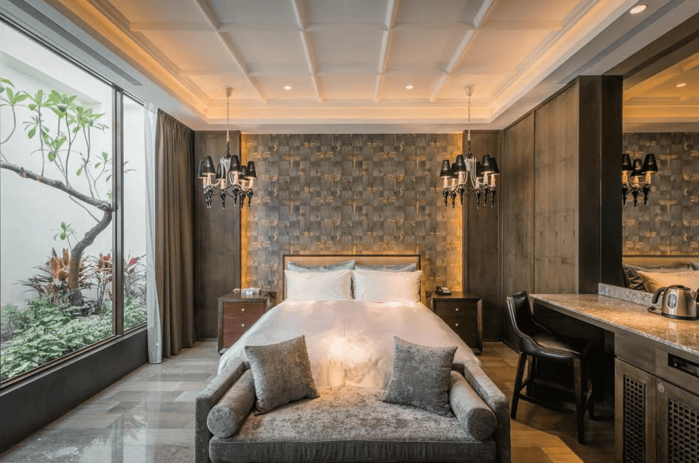 【員林住宿】棕櫚湖岸精品旅館的高級套房，房內擺設一張舒適的大床，搭配奢華的水晶吊燈與深色系牆面設計，營造低調奢華的氛圍。房間一側設有大片落地窗，能夠欣賞庭院綠景，並配有長沙發與大理石書桌，增添高級感。