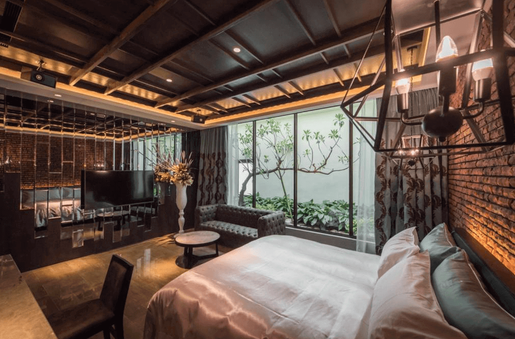 【員林住宿】棕櫚湖岸精品旅館的飯店客房，融合工業風與奢華元素，房內採用深色木質天花板與磚牆，搭配大型鐵製吊燈與鏡面裝飾。大床旁設有玻璃窗戶，可欣賞戶外庭院的綠意景觀，整體氛圍沉穩且浪漫。