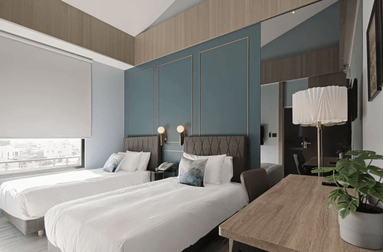 【台南安平住宿】森行旅的房間以墨藍與木質元素營造出現代簡約的高級感，配有兩張單人床與絎縫床頭板，床頭燈設計典雅，窗邊可享都市景致，另設有木質書桌與造型立燈，整體風格沉穩舒適，適合商務與旅遊入住。