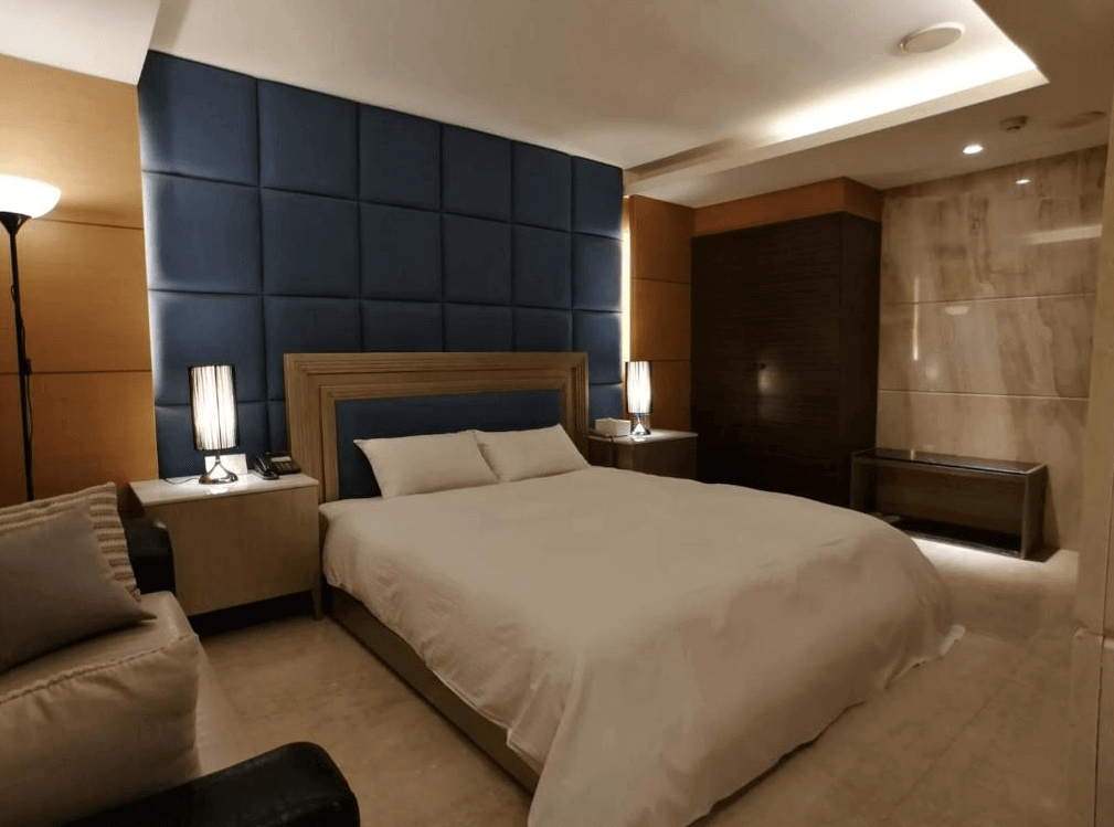 【員林住宿】楓華蜜雪兒精品旅館-員林館的房間，設有一張大床，床頭牆面採用深藍色絎縫設計，搭配溫暖的黃光床頭燈與大理石牆面，營造奢華感。房內擺放沙發與木質床頭櫃，整體色調沉穩優雅。