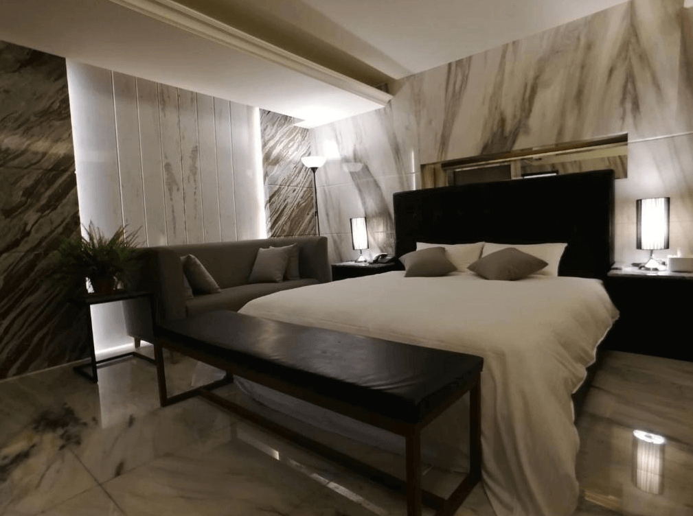 【員林住宿】楓華蜜雪兒精品旅館-員林館的高級飯店房間，以黑白大理石紋為設計主軸，床頭為黑色絎縫設計，搭配簡約的灰色沙發與黑色長凳，展現現代時尚風格。牆面間接燈光提升氛圍，空間寬敞舒適。
