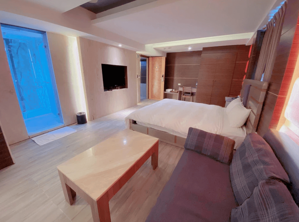 【員林住宿】楓華蜜雪兒精品旅館-員林館溫馨寬敞的飯店房型，設有大床、木質茶几與舒適沙發，營造居家感。房內配有大型壁掛電視，並設有玻璃隔間的藍光水療池，提供放鬆的住宿體驗。