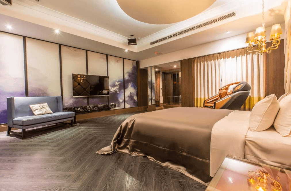 【員林住宿】楓采時尚汽車旅館的高級飯店套房，房間設有一張大床，搭配奢華的床頭燈與金色吊燈，營造典雅氛圍。牆面裝飾柔和雲彩圖案，增添藝術感。房內配有沙發、壁掛式電視，以及按摩椅，提升舒適度與放鬆體驗。