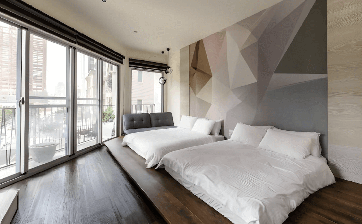 【台南安平住宿】樹屋文旅的現代簡約風格家庭房，設有兩張雙人床與一張灰色沙發床，房內以木質地板搭配幾何圖形設計牆面，大片落地窗可通往陽台，充滿都市景觀與自然光。