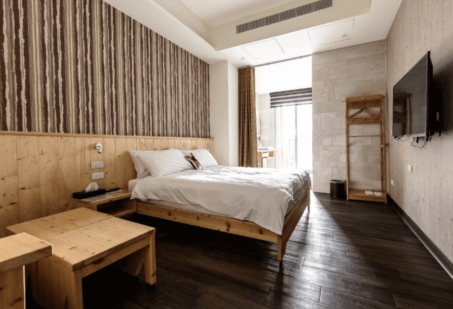 【台南安平住宿】樹屋文旅溫馨木質調客房設計，牆面採用木紋牆板與垂直條紋裝飾，搭配一張舒適的雙人床與簡約木作家具，空間寬敞且具備大片窗戶，營造出自然舒適的氛圍。