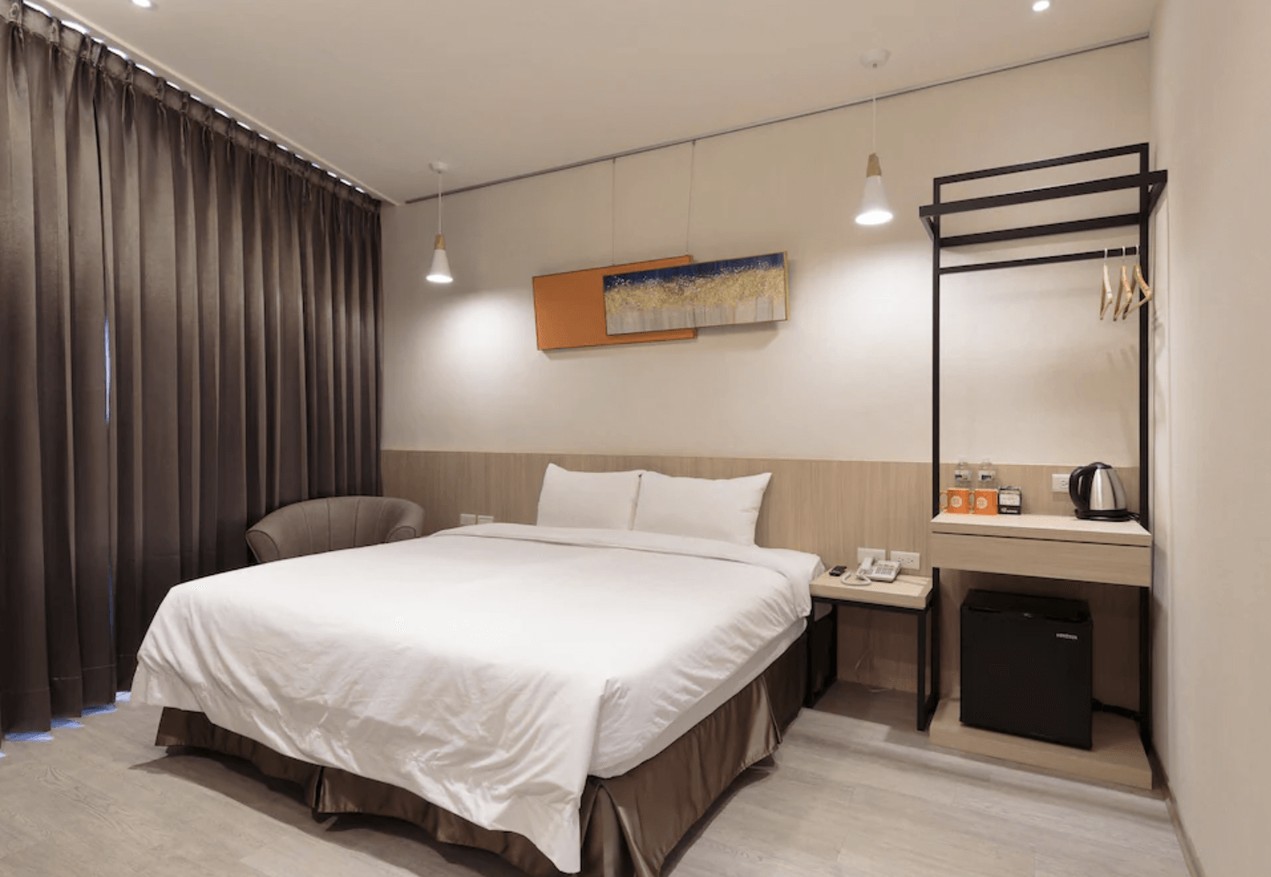 【小港住宿】橙屋商務旅館的雙人房，配有一張大床、咖啡桌椅組、開放式衣架與電熱水壺區。牆面上方掛有藝術畫作與簡約吊燈，室內採自然木色與米白色調，營造出清新現代的住宿氛圍。