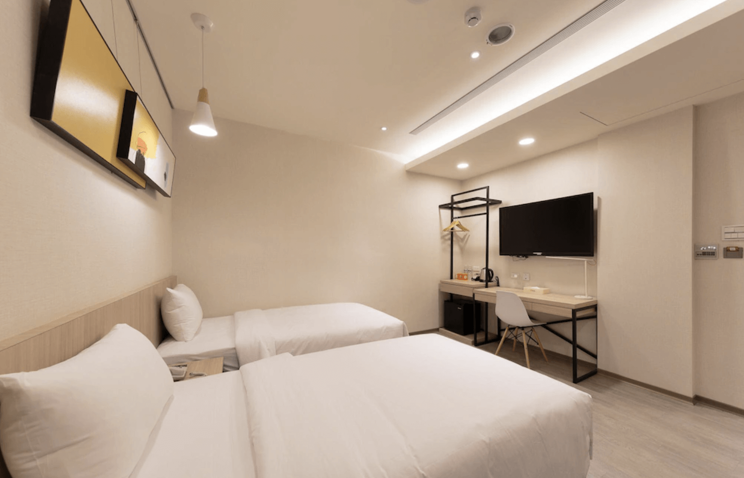 【小港住宿】橙屋商務旅館的現代簡約客房，牆上掛有平面電視，搭配書桌、吊燈、衣架與備品區，整體空間寬敞明亮，採用淺色木地板與米色牆面，給人整潔舒適的印象，適合朋友或家庭旅客。