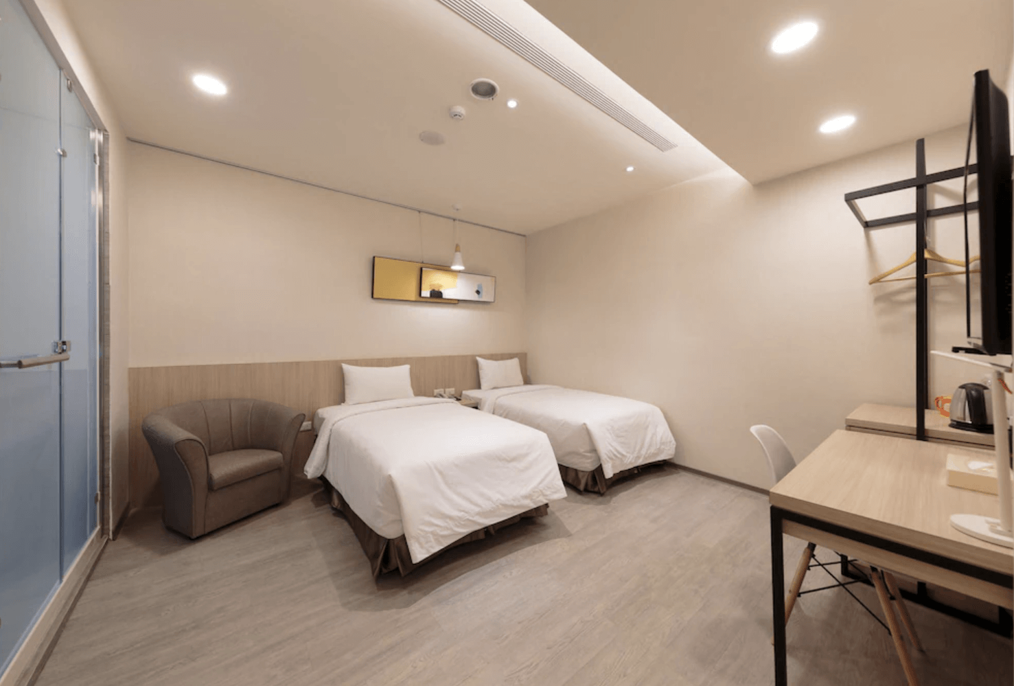 【小港住宿】橙屋商務旅館的雙床房，空間整潔寬敞，設有沙發椅、開放式衣架、電視與書桌，淺木地板搭配暖白燈光，使整體氛圍溫暖而不壓迫，是輕旅行或短期出差的不錯選擇。