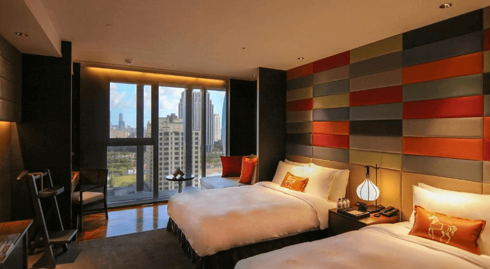 【高雄親子飯店】水京棧國際酒店時尚高級的雙床房，房內擁有大片落地窗，可欣賞都市景觀。牆面設計以多彩皮革拼接，營造現代藝術感。床鋪上擺放橘色抱枕，搭配典雅的燈具，呈現溫馨氛圍。房內配置木質家具與黑色元素，增添低調奢華感。