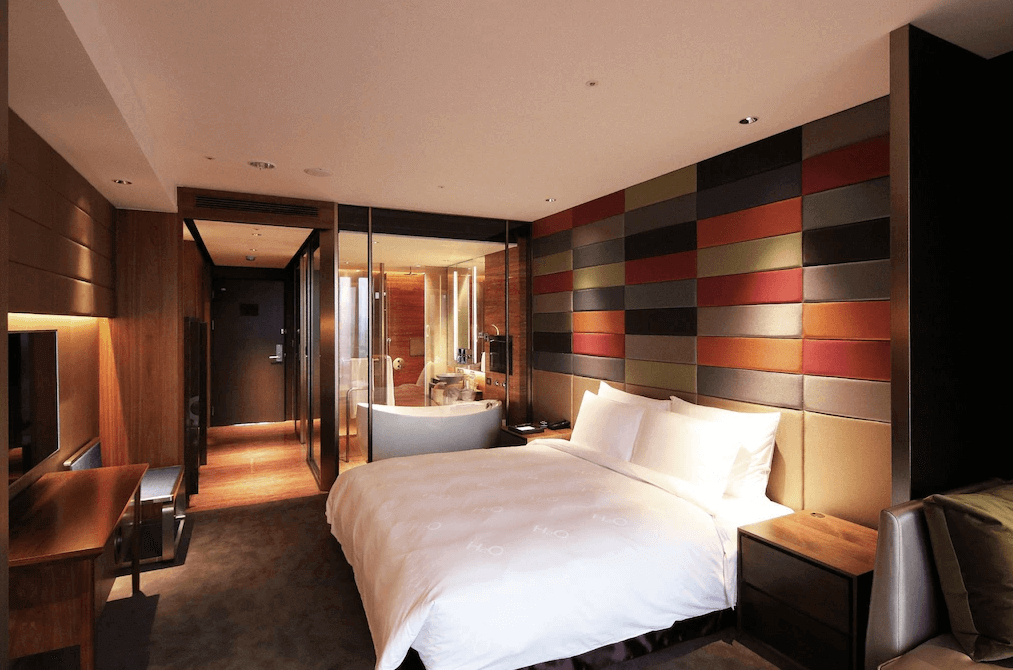 【高雄親子飯店】水京棧國際酒店的豪華雙人房，房內延續多彩皮革拼接牆設計，並採用木質調家具營造溫暖氛圍。寬敞的空間設計包含開放式浴室區，玻璃隔間內設有獨立浴缸與淋浴間，提升住宿的舒適感與便利性。