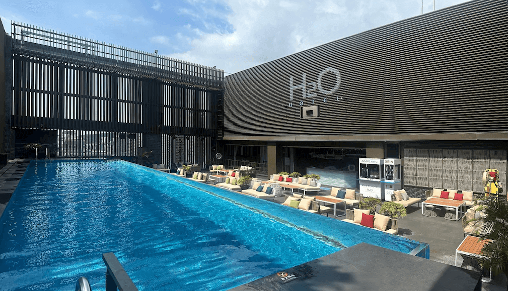 【高雄親子飯店】水京棧國際酒店的戶外無邊際泳池區，藍色泳池水面倒映著晴朗天空，周圍設有舒適的沙發座椅與抱枕，提供放鬆休憩的空間。背景為水京棧國際酒店（H2O HOTEL）建築外觀，設計融合木質百葉與現代風格，展現時尚度假氛圍。