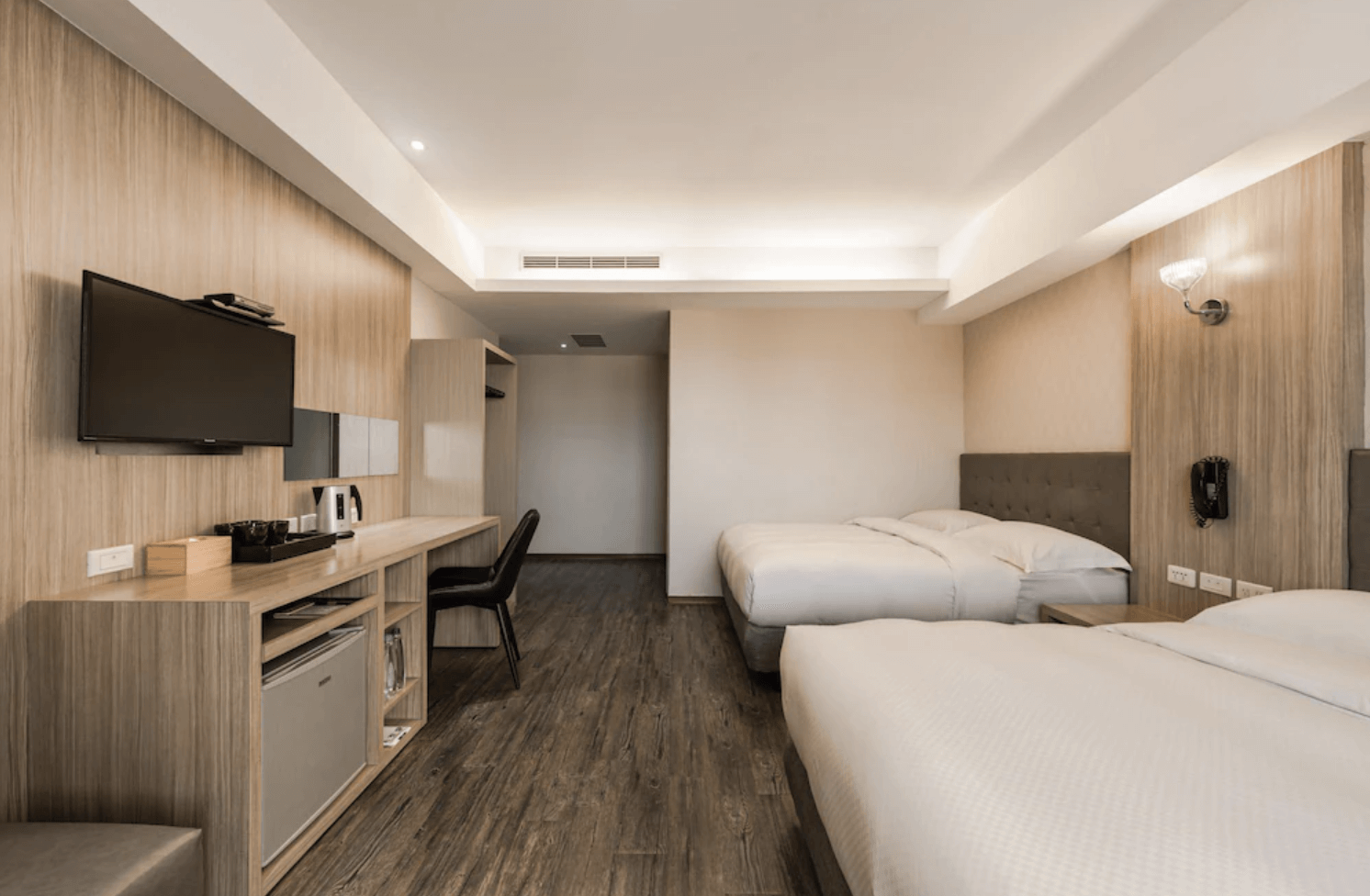 【小港住宿】河堤國際商旅的雙床房型，室內採用大量淺色木紋材質與深木地板，空間寬敞。房內設有兩張大床、平面電視、長型書桌與工作椅，並附有冰箱、熱水壺及茶具，整體感覺乾淨俐落，適合家庭或朋友同住。