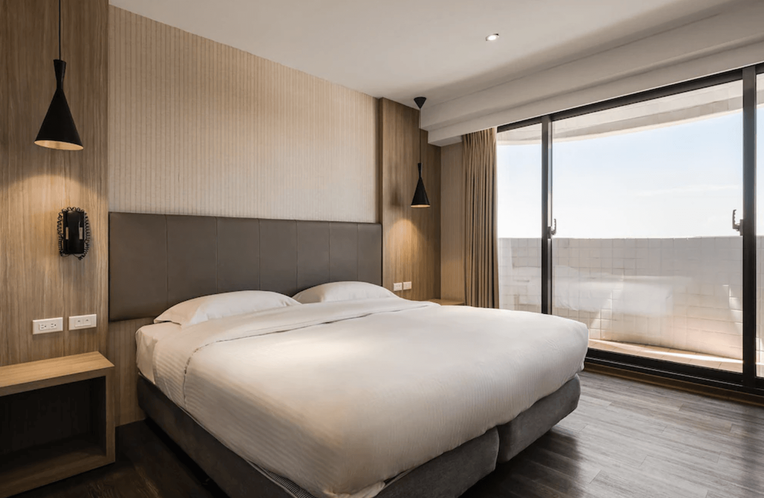 【小港住宿】河堤國際商旅的雙人房，設有一張大床與皮革床頭板，兩側設有簡約吊燈與插座牆，牆面為木紋設計，床邊是大片落地窗，外接陽台，光線充足。整體風格現代，適合商務或情侶入住。