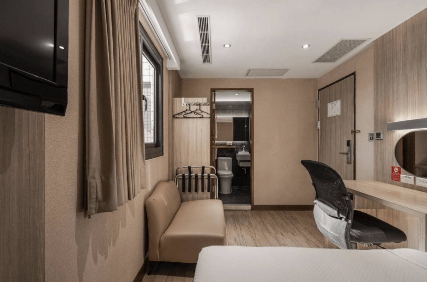 【小港住宿】河堤國際商旅的客房內部空間，畫面中可見電視、窗戶、沙發椅、書桌與人體工學椅，鏡子與燈光設計使空間更明亮。畫面遠方為浴室入口，可見馬桶與洗手台，整體布置簡約實用。