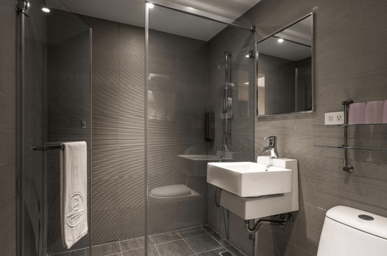 【小港住宿】河堤國際商旅的浴室，設計走現代簡潔風格，牆面與地板皆為深色紋理磁磚，配有玻璃乾濕分離淋浴間、簡約方形洗手台與馬桶，並有毛巾、備品與插座設計，空間整潔有質感。