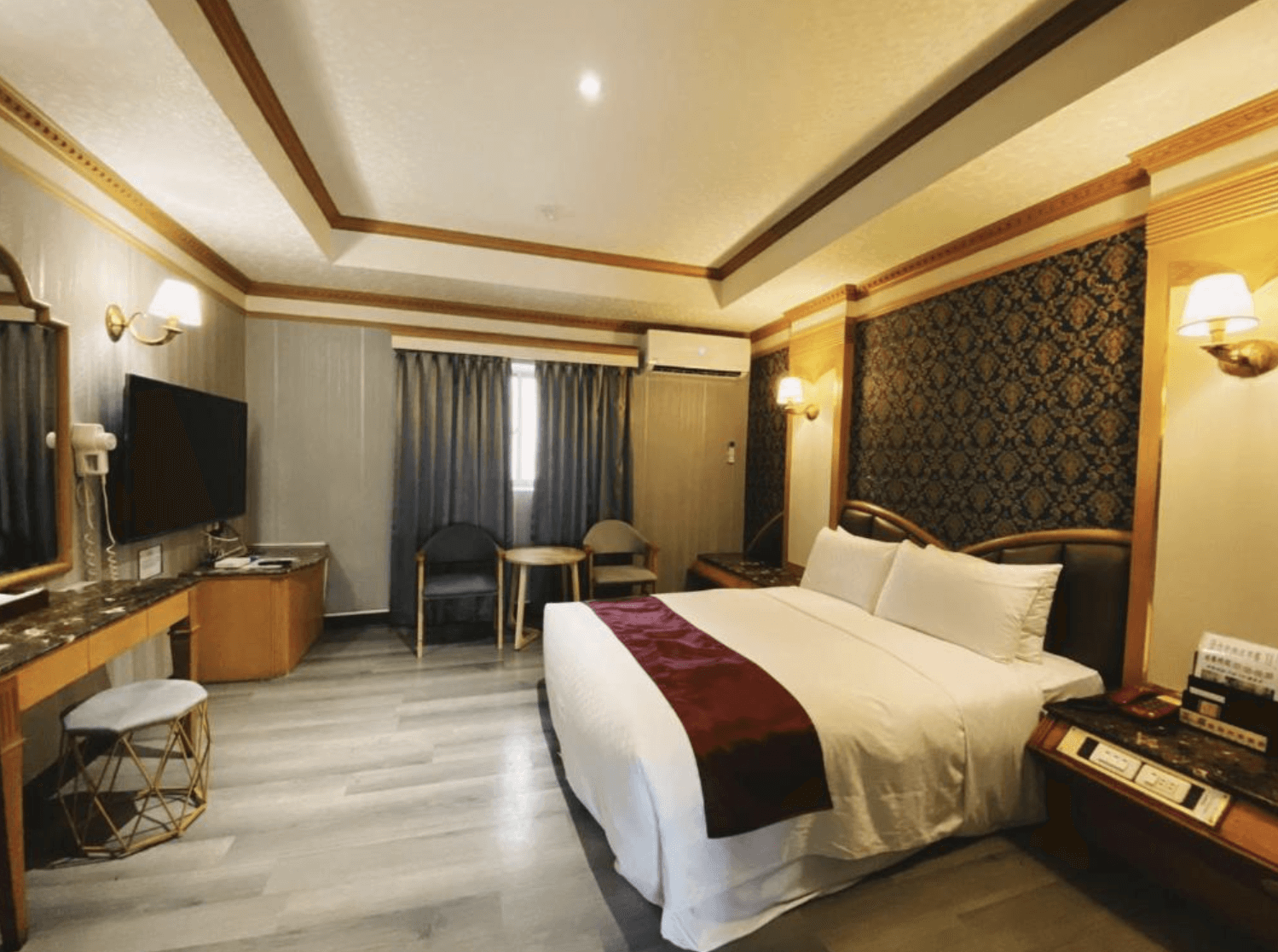 【小港住宿】艾萊汽車旅館具有歐式古典風格的客房，設有一張大床，床尾鋪有酒紅色緞面床旗。牆面以深色雕花壁紙與金色木框裝飾，搭配金邊壁燈及古典傢俱，營造出華麗典雅的住宿氛圍。房內配有電視、梳妝台與休憩桌椅，整體空間寬敞舒適。