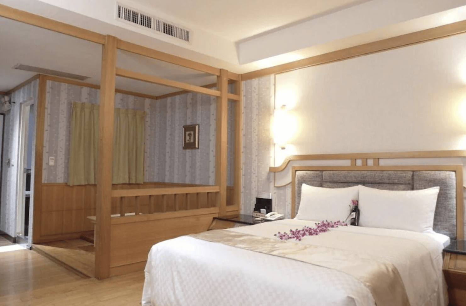 【小港住宿】艾萊汽車旅館的日式風格融合現代設計的客房，主牆為淺色系與木質線條構成，設有一張雙人床，床上擺放著香檳與紫色花瓣。左側為榻榻米木地平台區，可作為小茶區或休憩空間，整體氛圍溫馨放鬆。