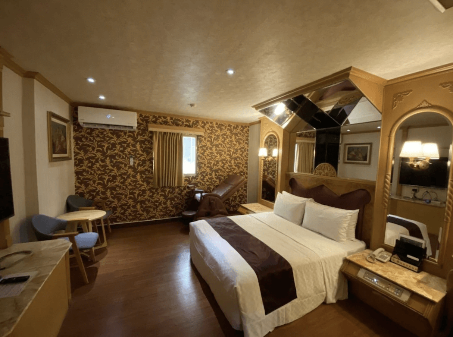 【小港住宿】艾萊汽車旅館的套房，房內配有大床、紫紅色床頭板與酒紅床旗，並搭配金色雕花牆飾與鏡面天花板。一側設有按摩椅與小圓桌椅組，牆面為繁複金色花紋，營造出豪華感受，適合情侶或特殊節日入住。