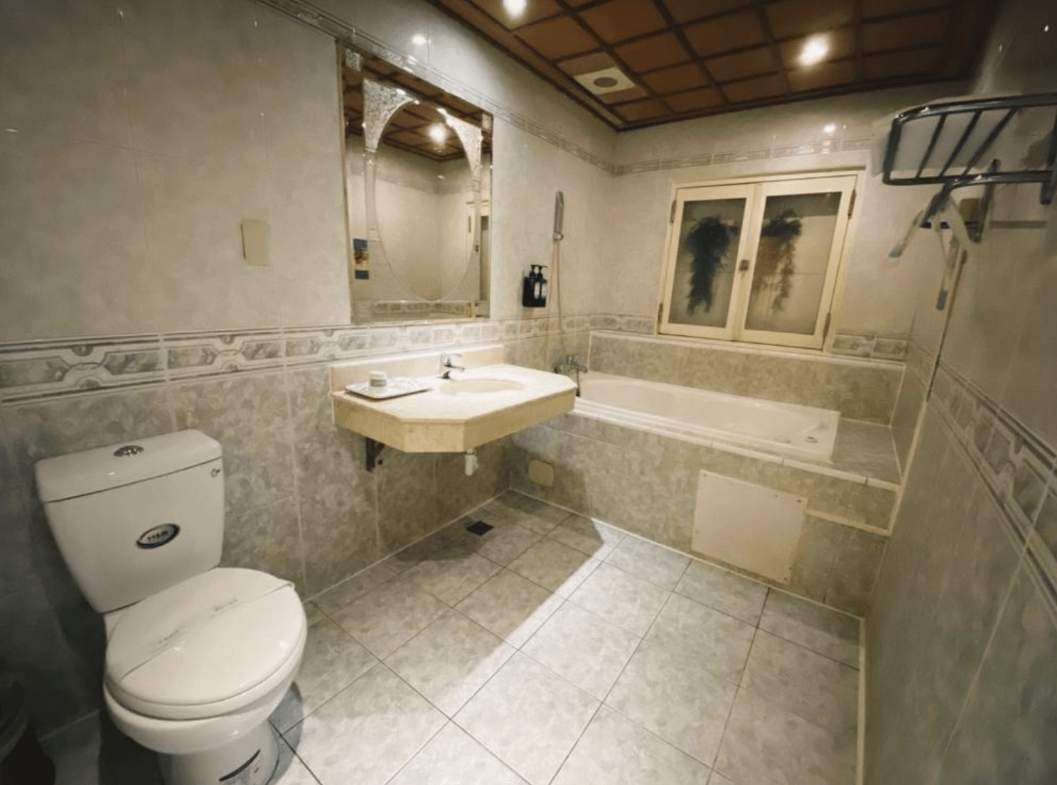 【小港住宿】艾萊汽車旅館的浴室空間，整體以米色磁磚為主，風格復古典雅。設有標準浴缸、洗手台與馬桶，鏡子上方有圓拱造型設計，天花板為木格紋裝飾，空間寬敞並提供基本盥洗用品，適合泡澡放鬆。