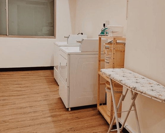 【斗六住宿】華安大飯店提供的自助洗衣房，設有洗衣機與烘衣機，旁邊擺放熨衣板、收納籃與清潔用品，空間整潔明亮，方便長住旅客使用。