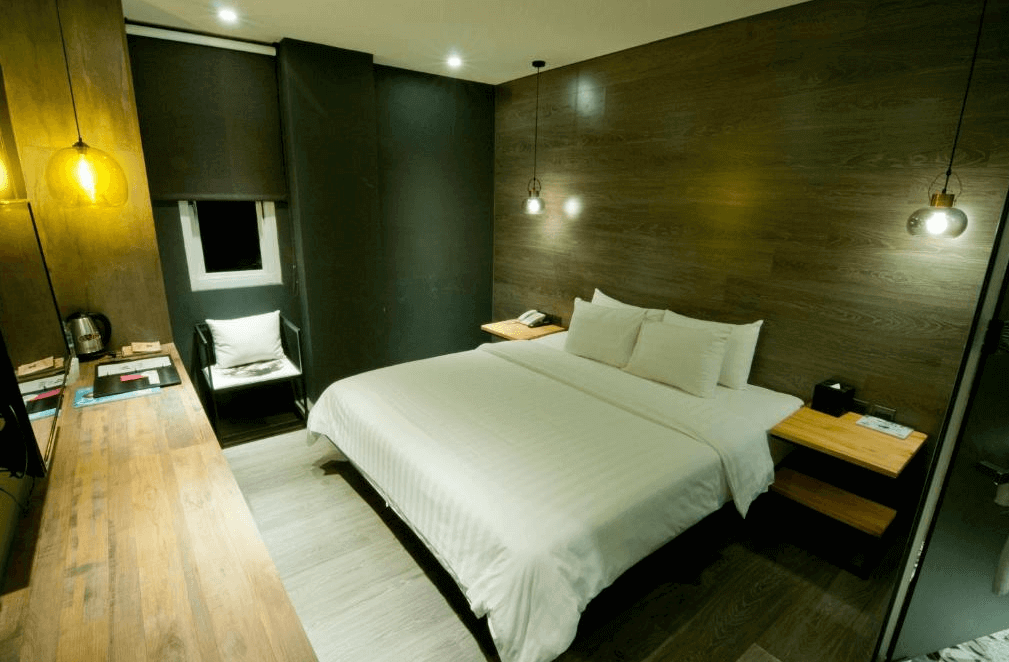 【虎尾住宿】虎尾春秋文創設計旅店的雙人客房，牆面以深色木紋設計，搭配溫暖的吊燈照明，白色大床整潔舒適，房內備有長型書桌、椅子與熱水壺，整體風格沉穩安靜。