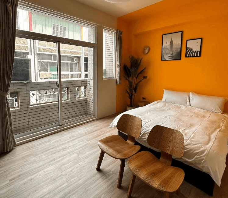 【虎尾住宿】虎尾漫遊的雙人房，搭配鮮橘色主牆與木質地板，落地窗讓自然光灑入室內，床尾擺放兩張木製椅子，空間明亮且富有設計感。