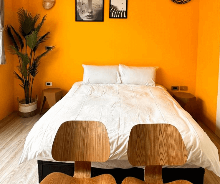 【虎尾住宿】虎尾漫遊的雙人床正面視角，鮮橘色牆面上懸掛兩幅黑白裝飾畫，床尾放置兩張造型木椅，角落植栽增添綠意，整體色調鮮明活潑。