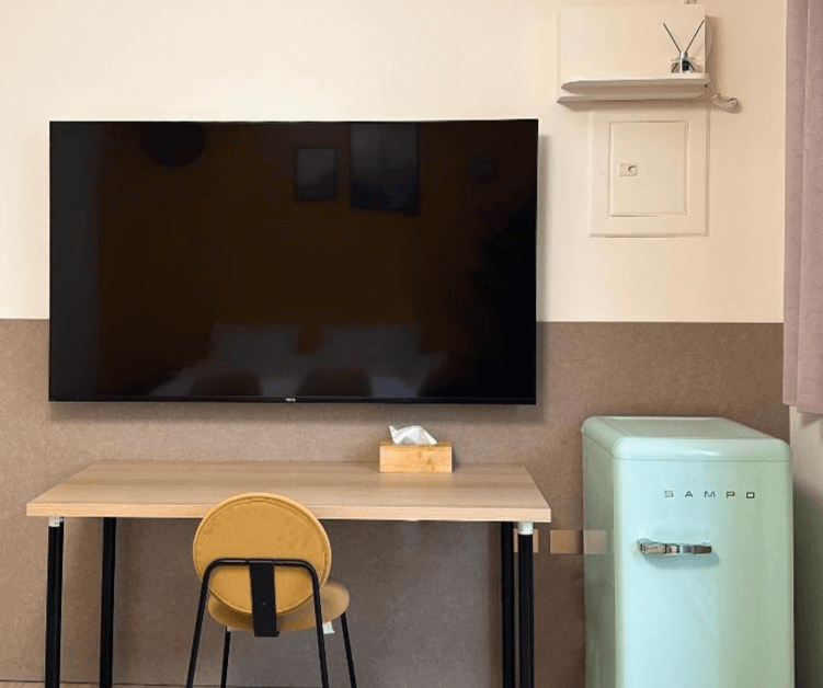 【虎尾住宿】虎尾漫遊的房內設有大型平面電視、木質書桌與圓背椅，旁邊為一台復古薄荷綠色迷你冰箱，整體色調溫潤，兼具復古與現代風格。