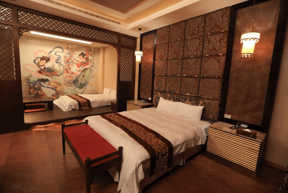 【斗六住宿】觀月商務休閒旅館的中式古典風格客房，主牆以立體紋理方格裝飾，床前擺放紅色坐凳，房內另一側有加大床與飛天仕女壁畫，融合傳統與現代設計，氛圍雅緻沉穩。