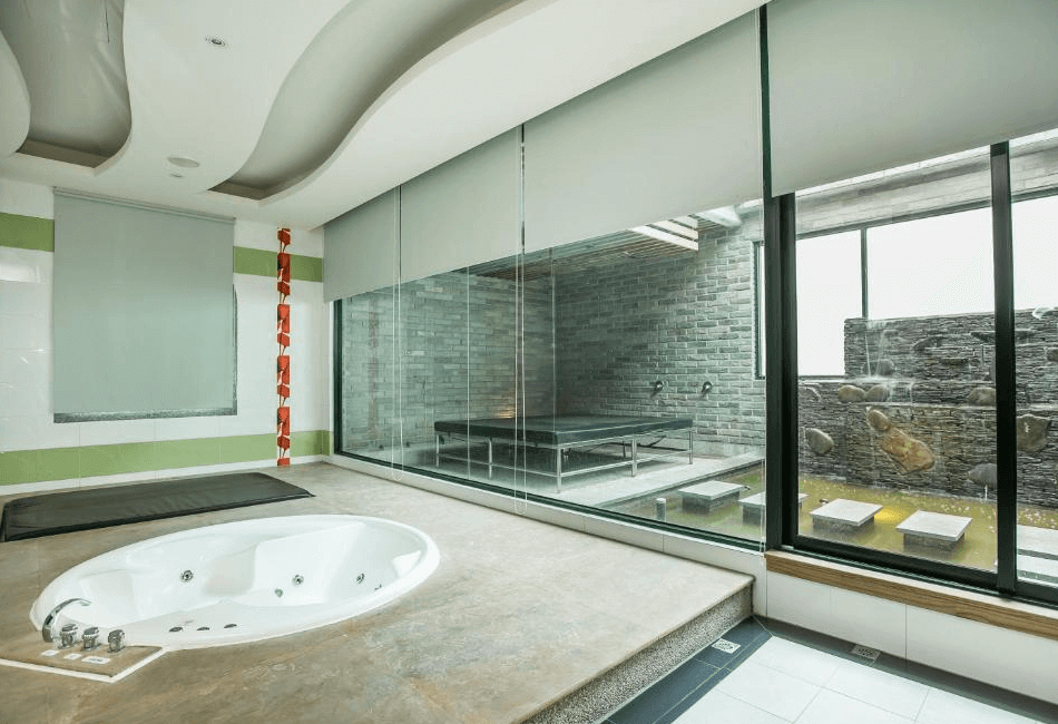 【斗六住宿】觀月商務休閒旅館的泡湯空間，設有一座嵌入式圓形按摩浴缸與地墊，大片落地窗外可見戶外造景區，有石牆流水與小型水池，營造出日式風呂的靜謐與自然氛圍。