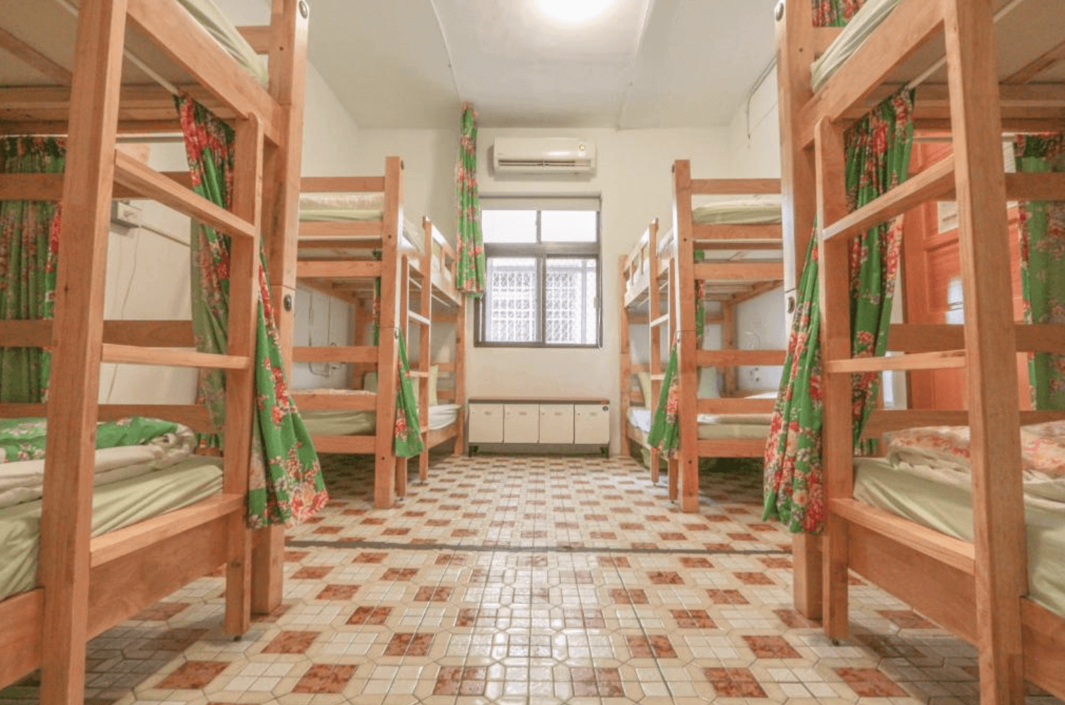 【台中背包客棧】踢‧生活背包客棧的宿舍房,房內擺放了六張上下鋪木製床架,每張床鋪配有綠色碎花窗簾提供隱私。地板鋪有復古風格的磁磚,房間中央留有寬敞的通道,並設有空調與窗戶,整體呈現懷舊風格。