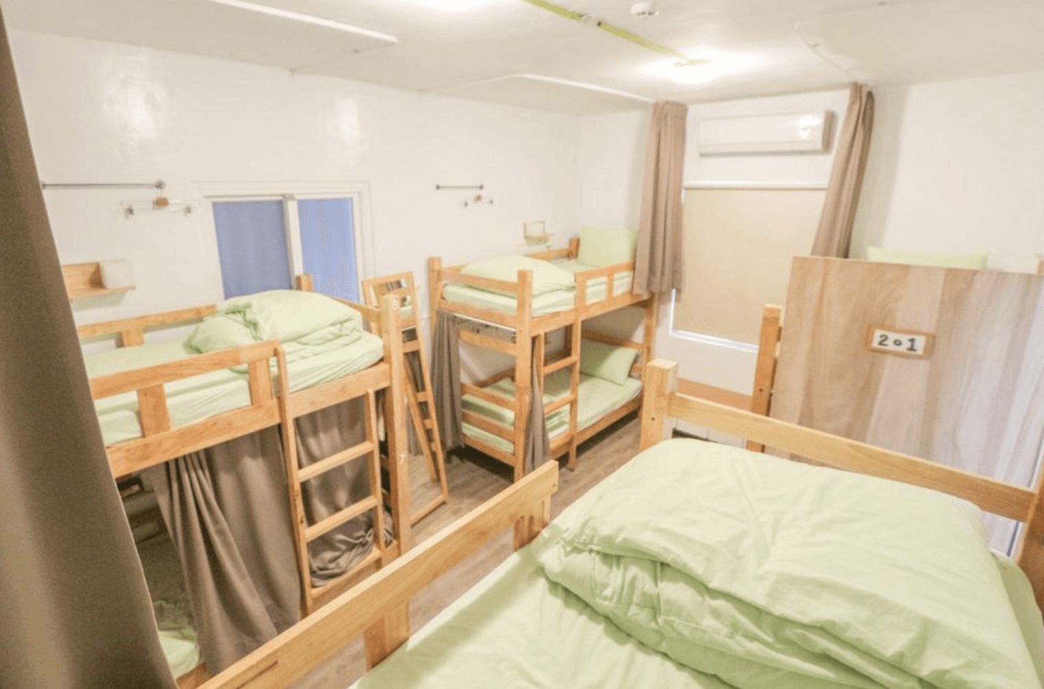 【台中背包客棧】踢‧生活背包客棧的宿舍房,採用木製上下鋪床架,床鋪搭配淺綠色寢具與米色窗簾,每個床位皆配有獨立遮簾增加隱私。房內設有空調與大窗戶,整體環境明亮整潔,地板為淺木紋設計,牆面為白色簡約風格。