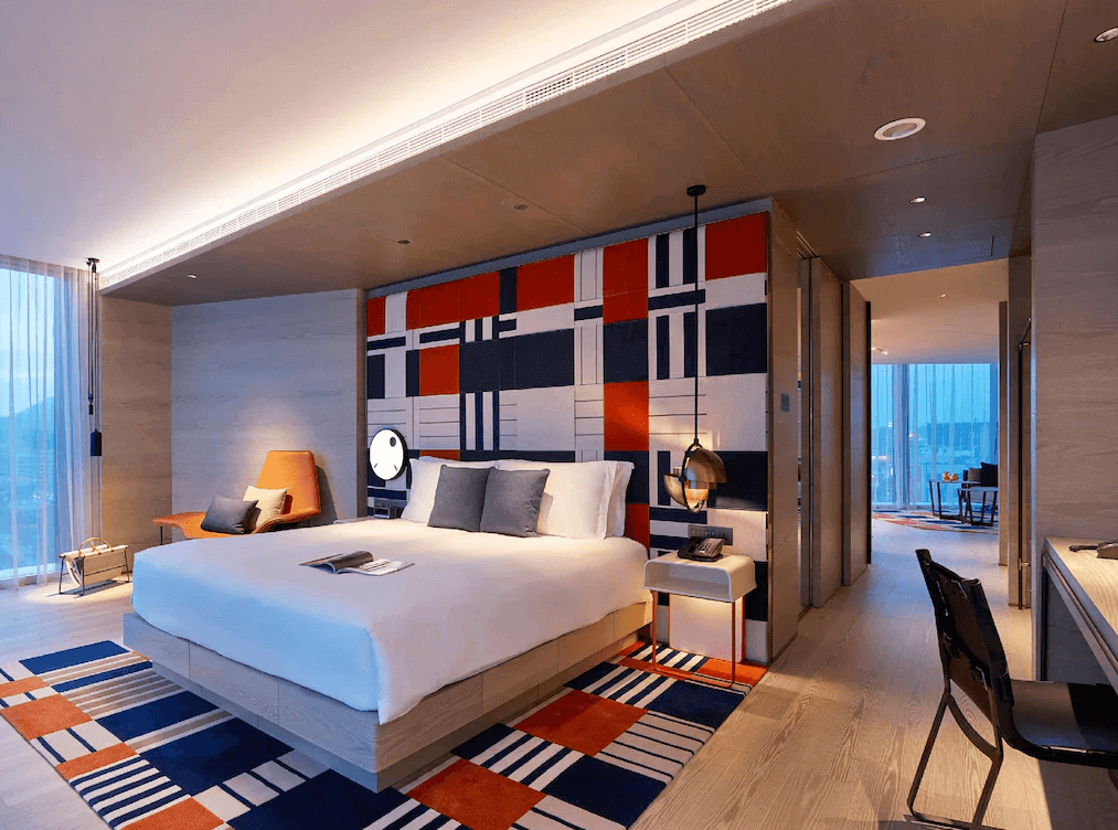 【高雄親子飯店】"高雄寵物友善住宿"“高雄中央公園住宿”高雄中央公園英迪格酒店時尚藝術感的豪華雙人房，房內設計以幾何圖案為主題，床頭牆面與地毯融合橘、藍、白的現代風格元素。房內設有舒適的大床、橘色休閒椅、懸吊式床頭燈，以及通往客廳的開放式空間，搭配大片落地窗，可欣賞城市景觀。