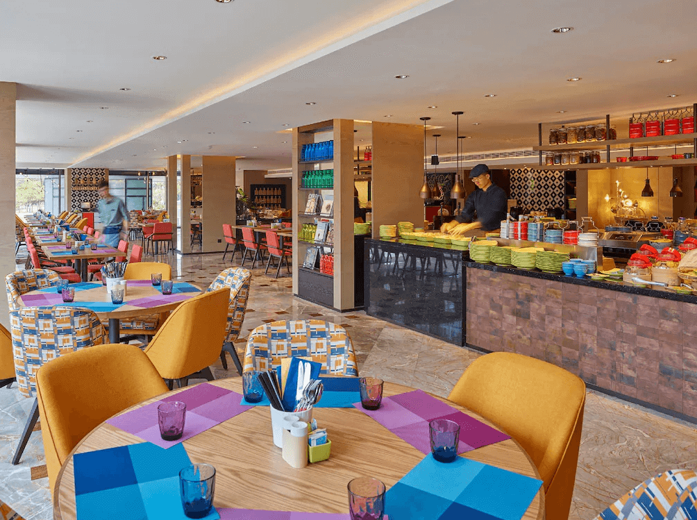 【高雄親子飯店】"高雄中央公園英迪格酒店精選示意圖"高雄中央公園英迪格酒店充滿活力與色彩的飯店餐廳，採用鮮明的黃色、藍色與橘色家具，營造熱鬧氛圍。開放式廚房提供多種現場料理，擺放整齊的餐具與裝飾品增添用餐樂趣。整體設計結合時尚與工業風，打造獨特的用餐體驗。