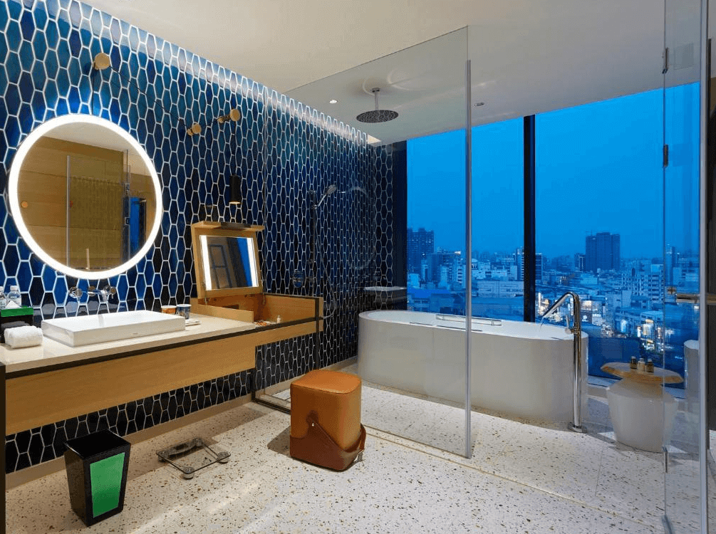 【高雄親子飯店】"高雄寵物友善住宿"高雄中央公園英迪格酒店的高級景觀浴室，設計以藍色蜂巢狀磁磚與玻璃隔間為主，呈現摩登質感。浴室內設有獨立浴缸，可一邊泡澡一邊欣賞城市夜景，並配有圓形LED燈鏡、梳妝台與現代化洗手台，提供頂級舒適的洗浴體驗。